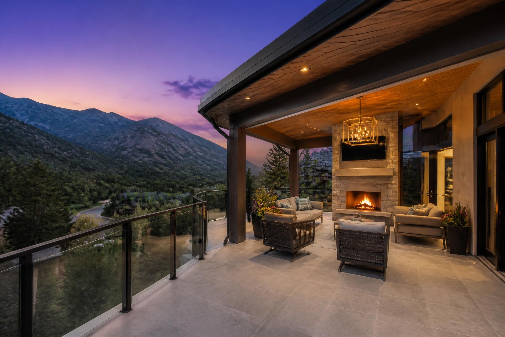 Covered outdoor living room with fireplace and glass railing at a custom home in Cottonwood Heights, Utah at dusk with canyon views