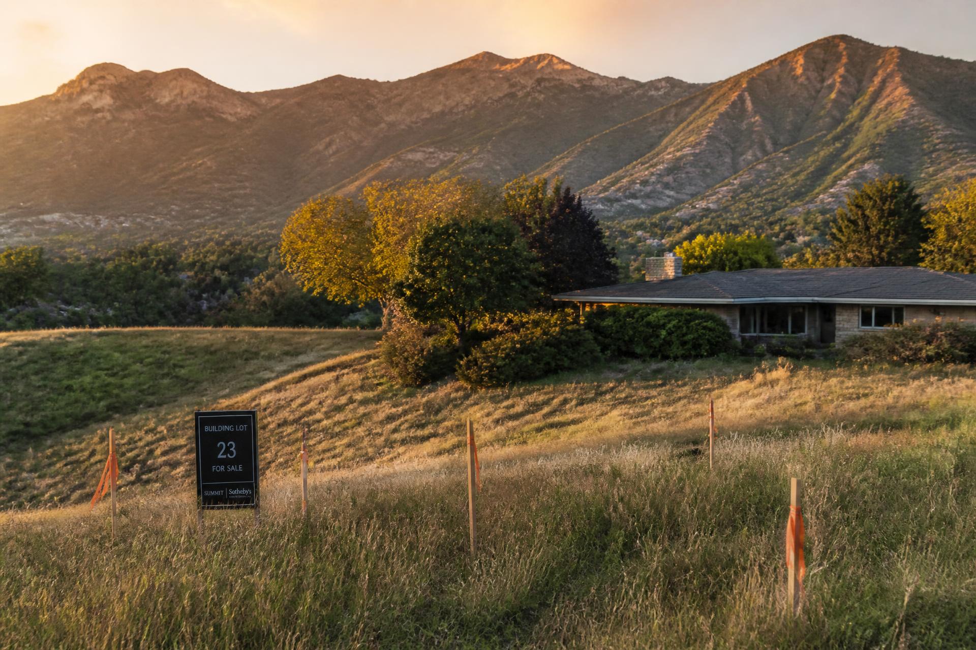 Vacant hillside building lot with survey stakes and flagging beside an older ranch home in the Salt Lake Valley foothills at sunset